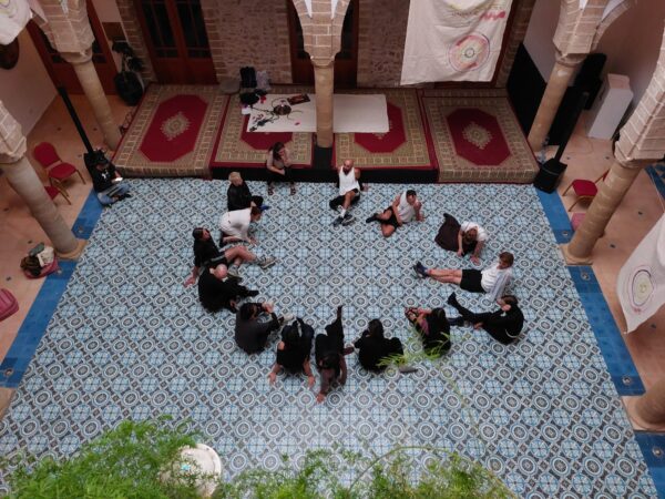 A group of people sits in a half-circle on a blue-and-white patterned tiled floor in a historic space with stone columns and carpets along the walls; painted cloths hang above.