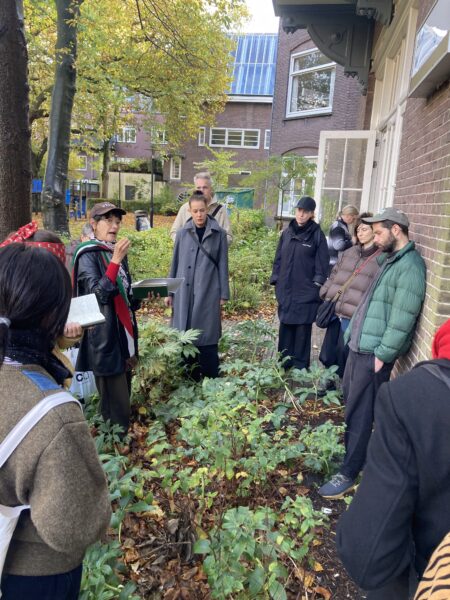 Artist Milena Bonilla is standing knee-high in vegetation and is speaking to a small group of gathered people, some of which are leaning on a brick building of the WG-terrein in Amsterdam West.