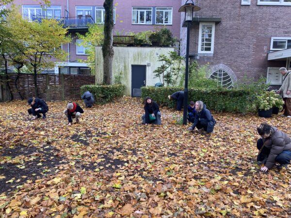A small group of people are seen scattered, kneeling and touching the ground which is covered in yellow, brown and green leaves. They are surrounded by the brick buildings and trees of the WG-terrein in Amsterdam West.