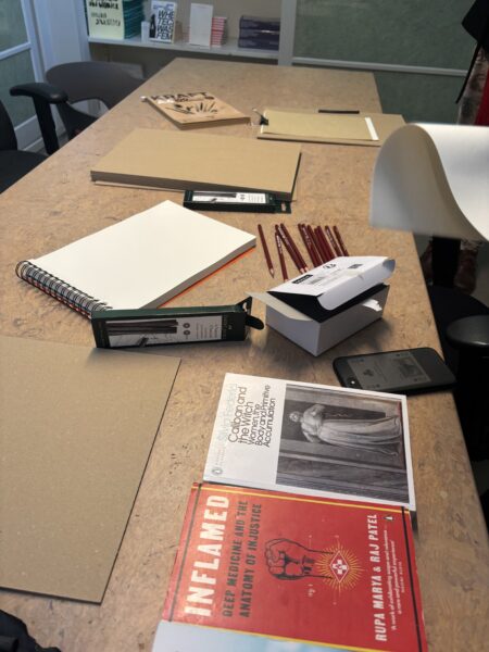 A working table on which are scattered drawing materials such as paper, charcoal pencils and clip-boards. Two books are also seen, one with a red cover with the title ‘Inflamed: Deep Medecine and the Anatomy of Injustice’; the other with a white cover with the title ‘Caliban and the Witch: Women the Body and Primitive Accumulation’.
