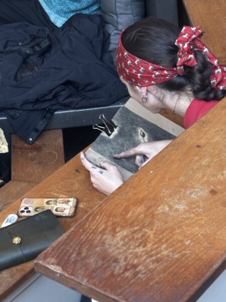 A woman, with a red headscarf in her long straight brown hair, is seen from the back, seated in the wooden tribune of the Collegezaal of the WG-Terrein. She is bent over a clipboard, and is busy working on a charcoal drawing of a flower.