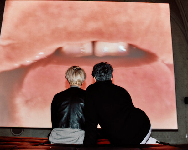 Two people sitting on a bench. They are seen from the back and are facing a large screen on which a close-up of moist lips pressed up against a glass surface dwarfs their bodies.