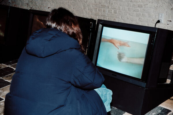 A person wearing a dark blue winter coat is sitting facing a TV set in a large black incasing on the floor. On the screen is an image of two arms, visibly belonging to two different bodies, whose hands reach out toward each other in a body of light blue water.