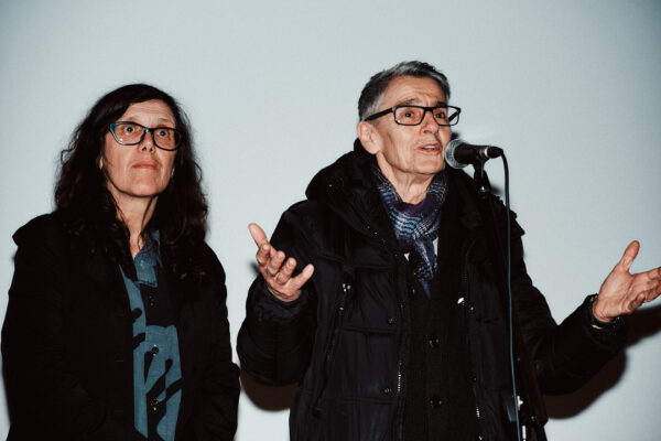 Artists Christina Della Giustina and Anchan/Anna Daučíková are seen in a close-up. Anchan/Anna Daučíková has her arms raised and is speaking into a mic, while Christina Della Giustina, stands slightly behind looking into the distance.