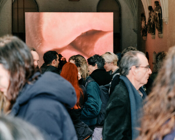 A crowd of people engaged in conversations throughout the hall of the cavernous Gothic architecture of the Vleeshal in Middelburg. Above their heads towers the projected image a close-up of a pair of lips.