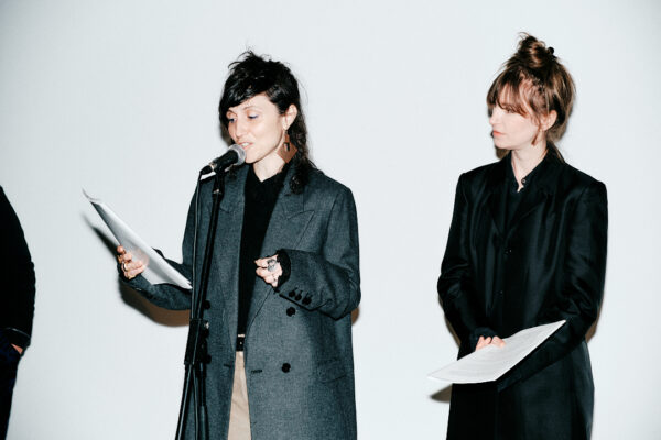 Curators Sara Giannini and Martha Jager are seen close-up. Sara is looking downward at sheets of white paper she holds in her right hand while speaking in a mic, while Martha stands to her right, with an inclined head, listening, while also holding sheets of white paper.