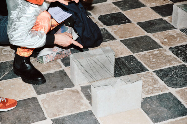Two bodies are crouched around a small sculpture, their hands pointing to a small sculpture made of concrete blocks and shards of glass on the floor.