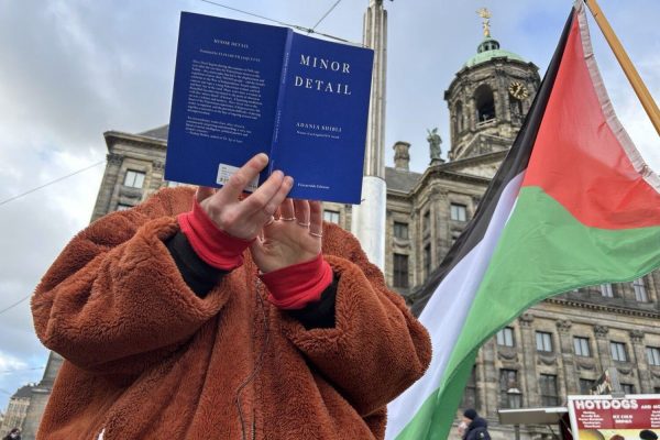 A shot looking upward at a person wearing a fuzzy brown fleece, holding a blue book open so that we cannot see their face. The words Minor Detail by Adania Shibli are written in white on its cover. To the right of the person is a Palestinian flag blowing in the wind and behind one can see the tower of the palace on the Dam Square of Amsterdam. Above is a bright blue sky.
