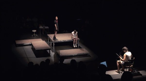 A dark theatre space viewed from the top of the audience riser. In the centre of the space, a spotlight is set on five bare stage parts. An actress in a short blue dress, and braided hair stands on the left, looking over her shoulder. An actress in a short white dress sits on the edge of the stage, her legs dangling above the theatre floor, looking down. She holds a computer mouse in her hand. Opposite the actresses, in the bottom right of the image, a woman in a traditional white vest, seated on a wooden café-style chair, looks down at her instrument as she plays the lyra and bow.