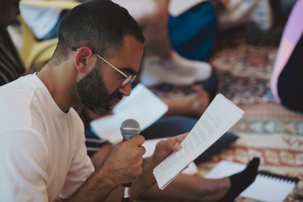 A person wearing glasses holding a mic and a script up to their face while they read.